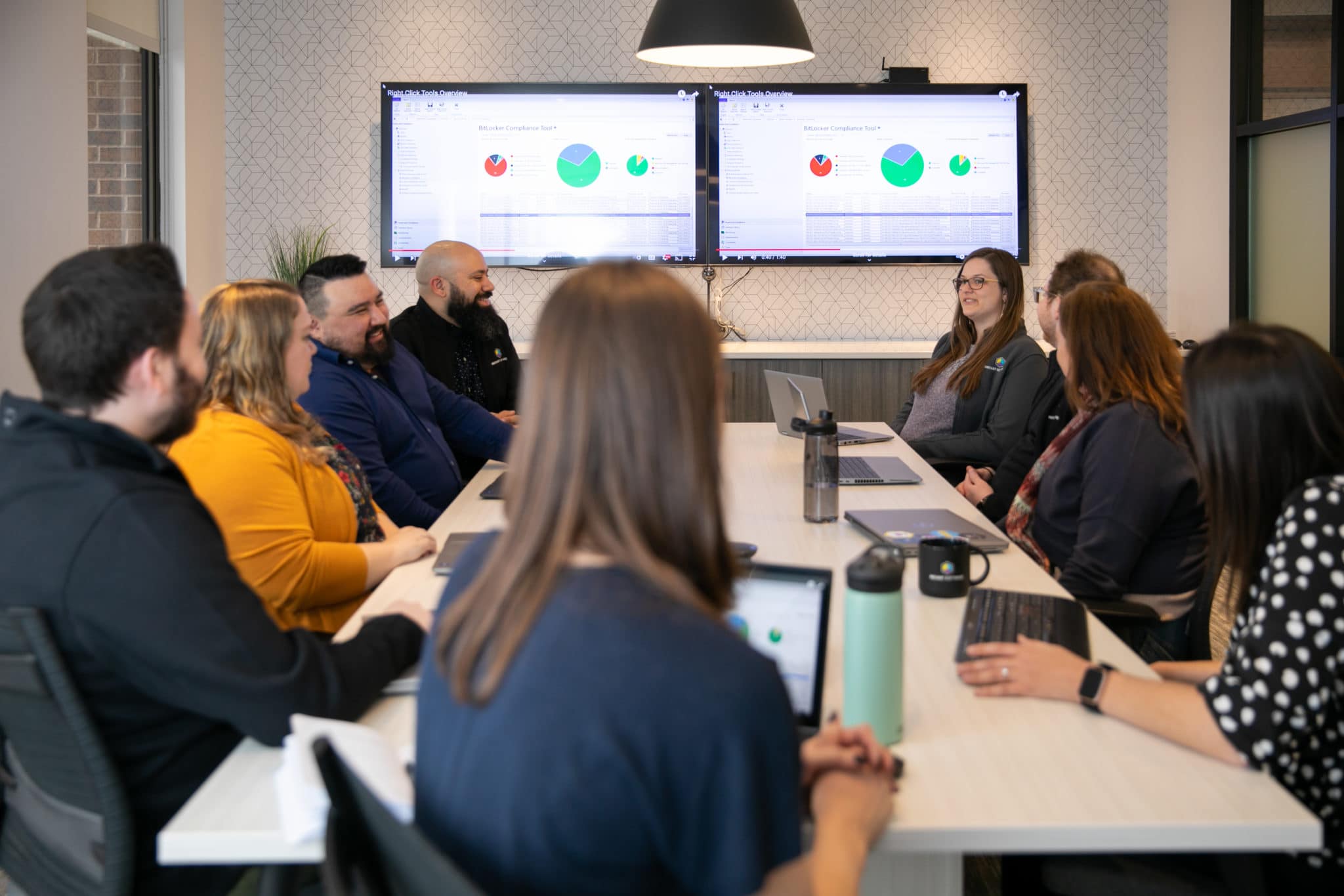 Table surrounded by sitting Recast Software employees that are meeting and reviewing Right Click Tools reports on two large monitors.