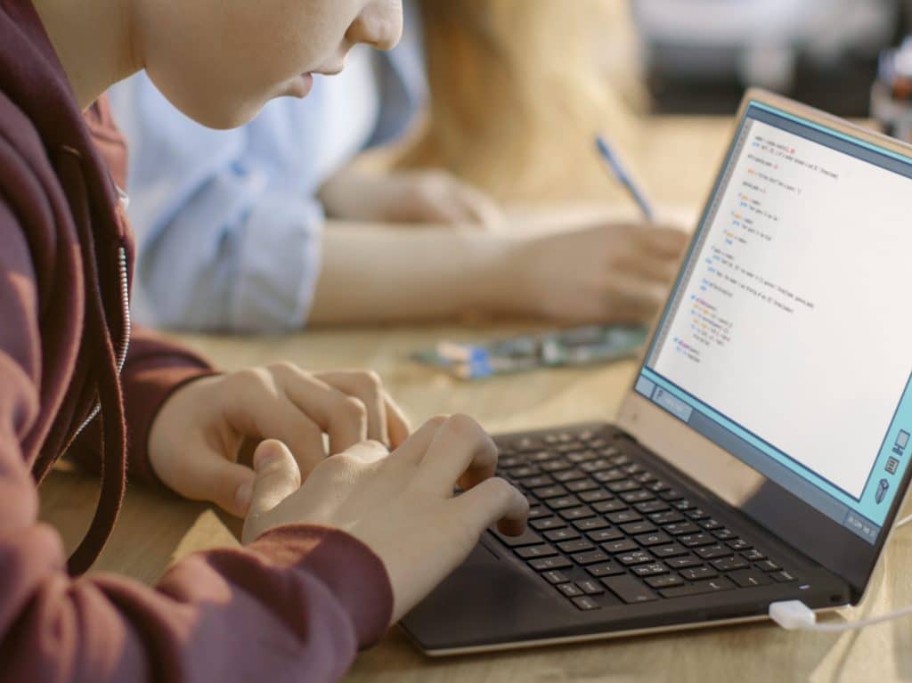 A child working on a computer doing schoolwork. Other kids' seen in the background.