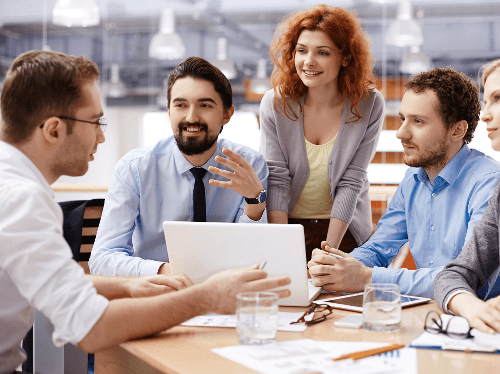 A professional setting with a staff of five meeting with paperwork of graphs around them with also their laptops and tablets.