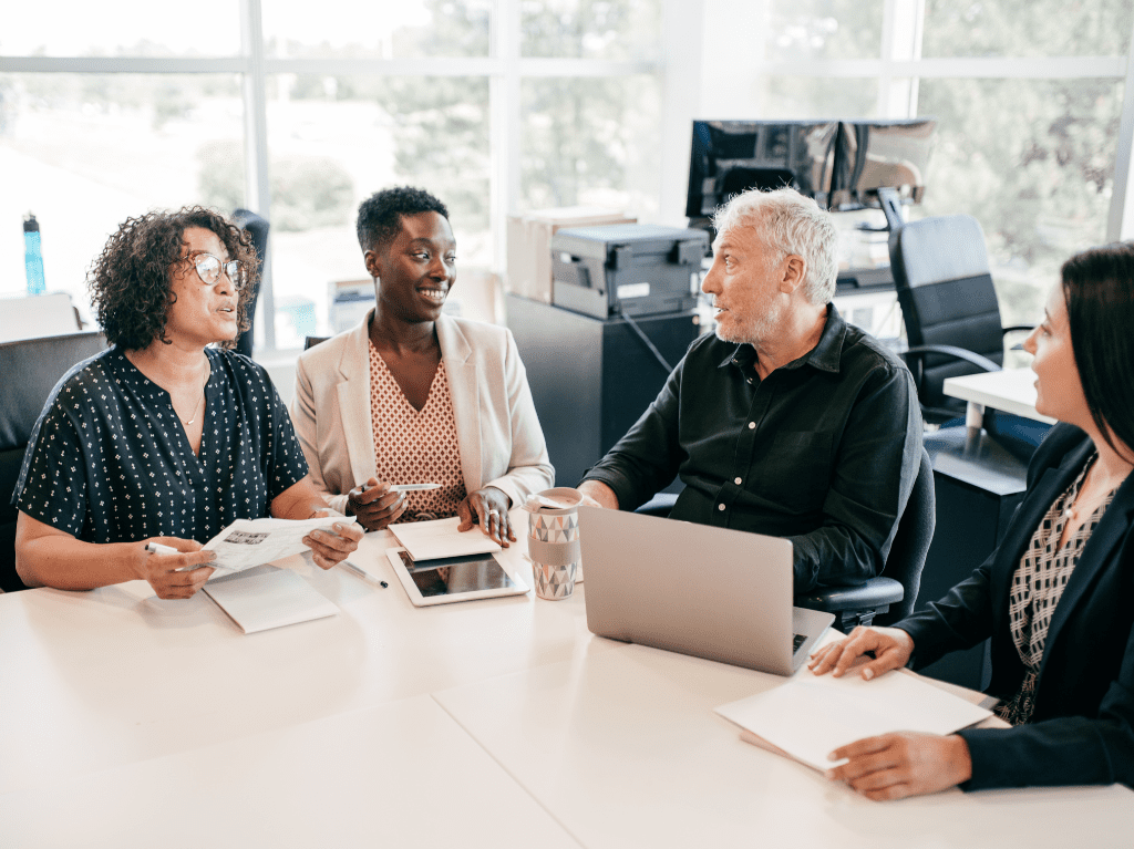 A group in an office setting sitting around a table in a meeting and using various devices like laptop and tablets.