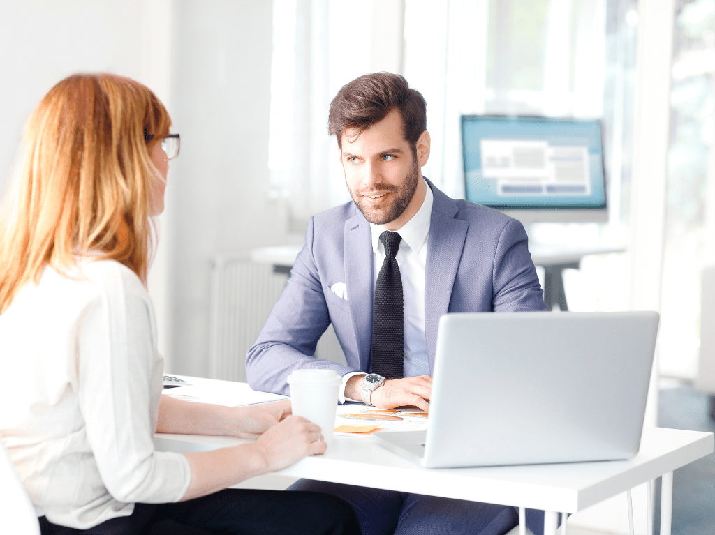 Man and woman meeting at a desk while he is using a laptop.